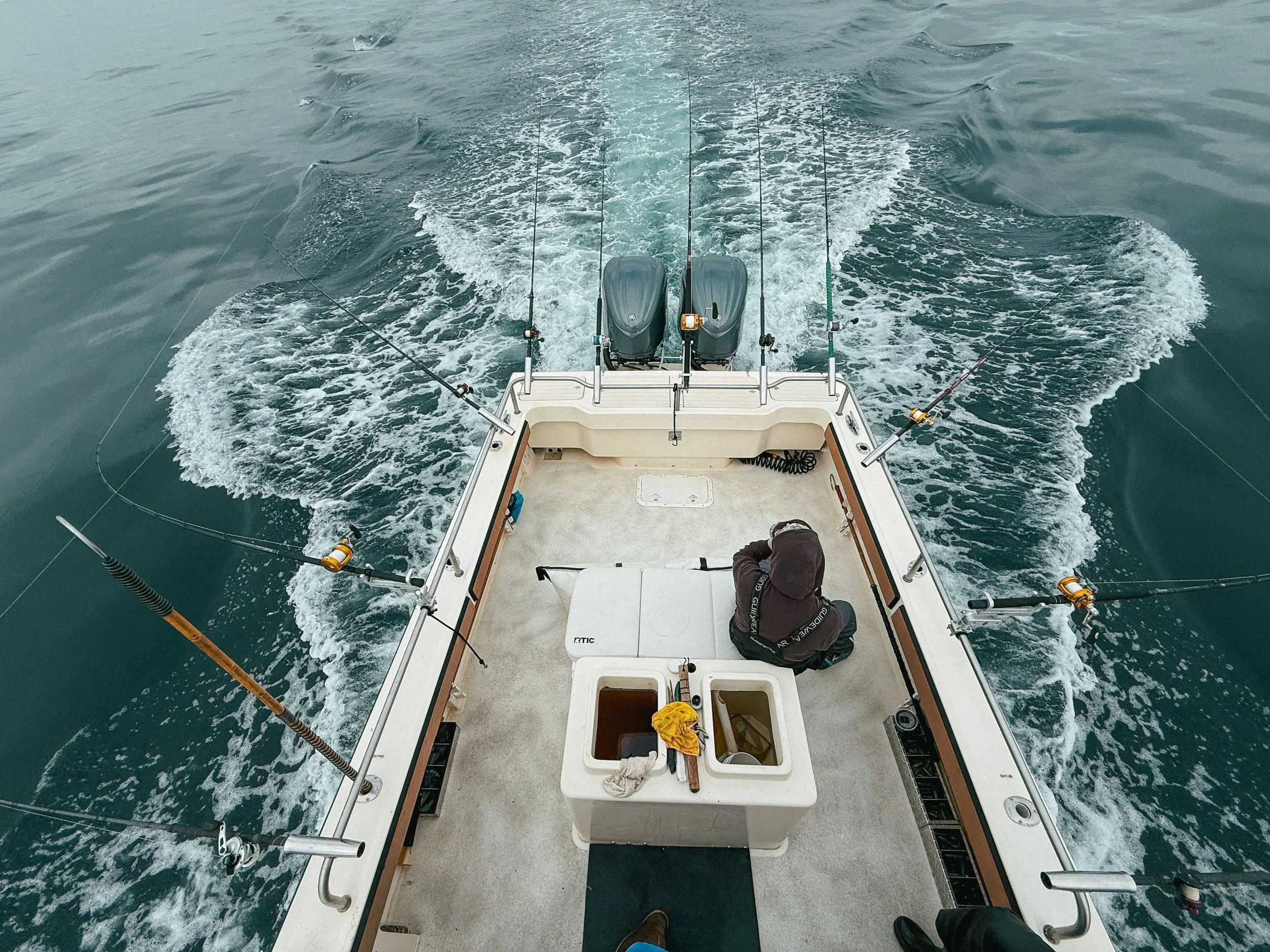 A person sitting on a fishing boat in the middle of the water with fishing rods attached to the sides, and two outboard motors at the stern, creating a wake behind the boat.