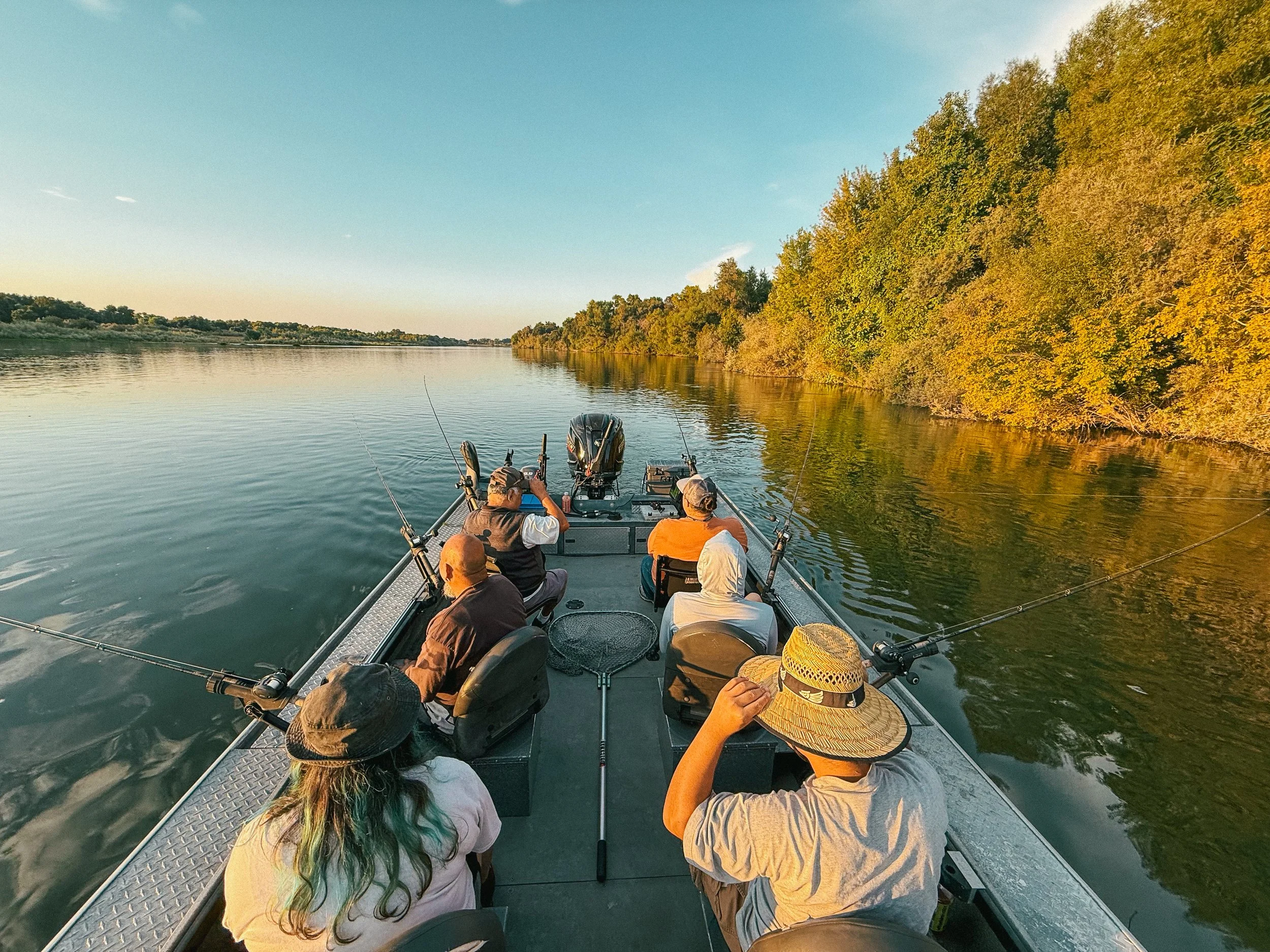 A group of people fishing from a boat on a calm river during sunset, with trees along the shoreline.