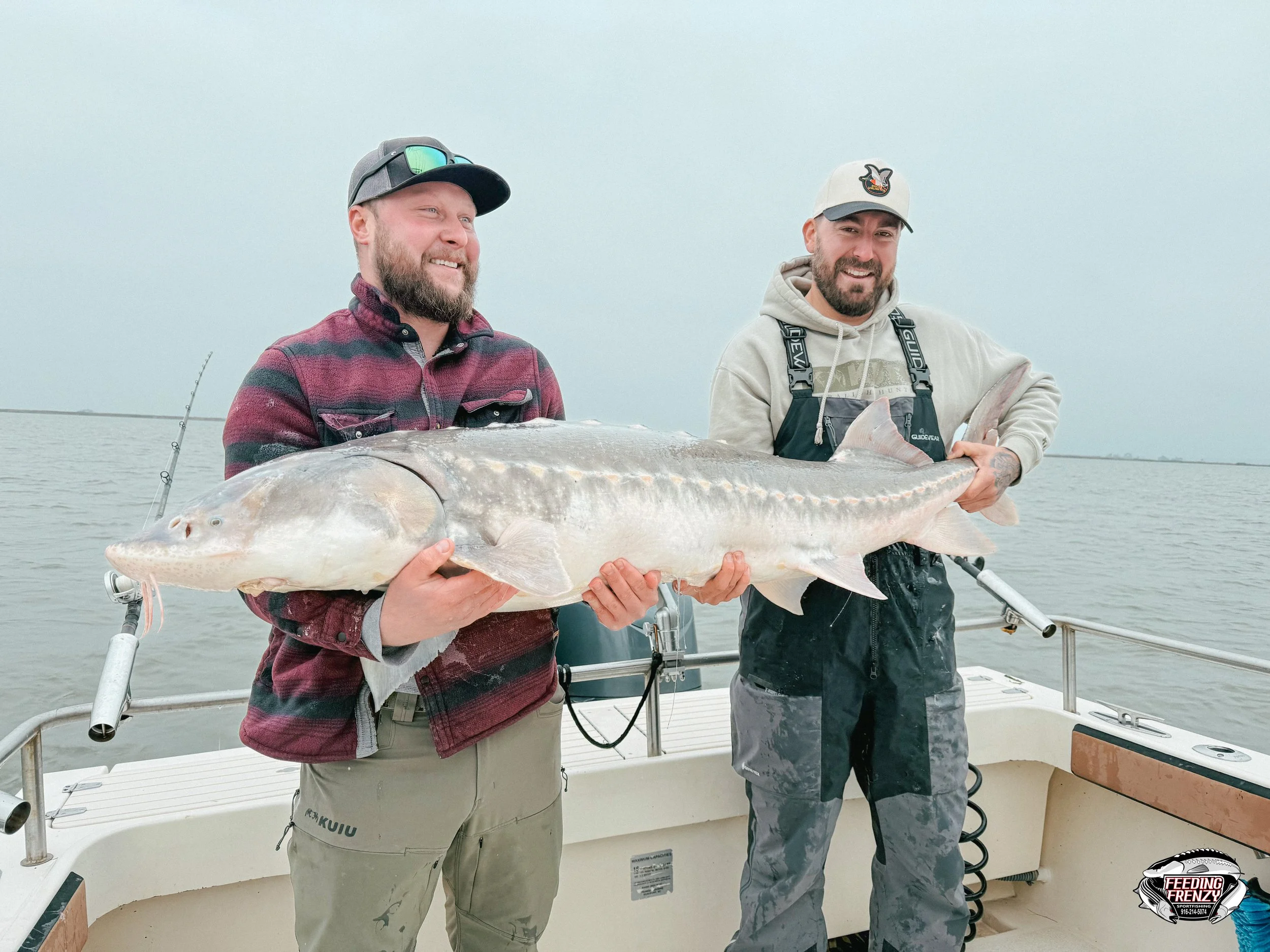 Two men standing on a boat holding a large fish together, smiling at the camera, with water in the background.