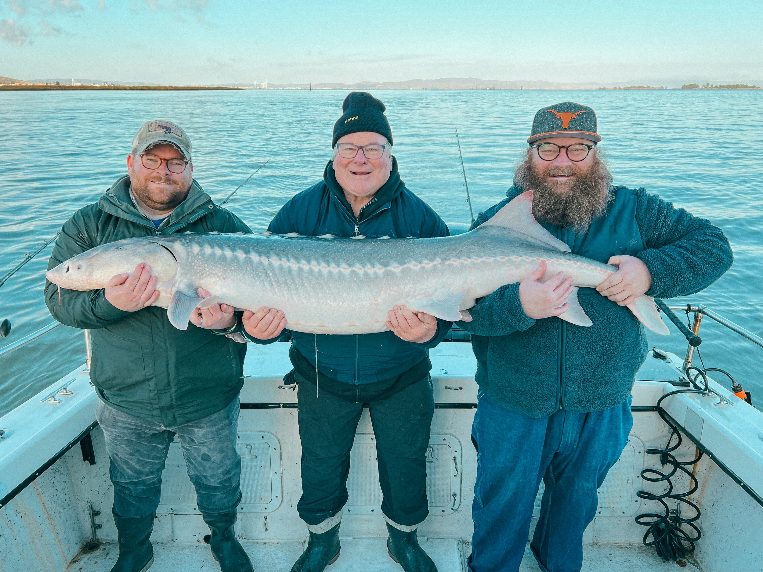 Three men on a boat holding a large fish, likely a sturgeon, with a body of water and distant shoreline in the background.