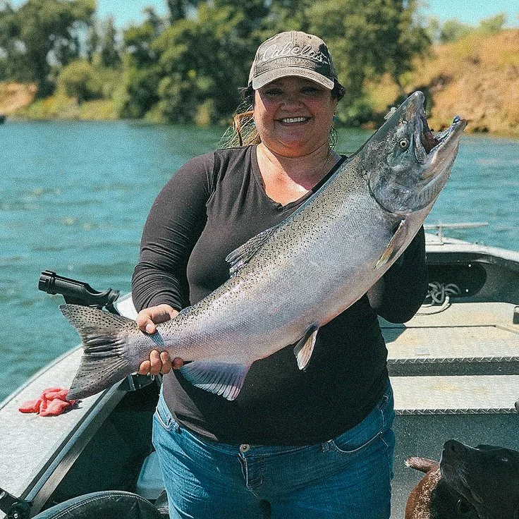 A woman smiling and holding a large fish on a boat with water and trees in the background.