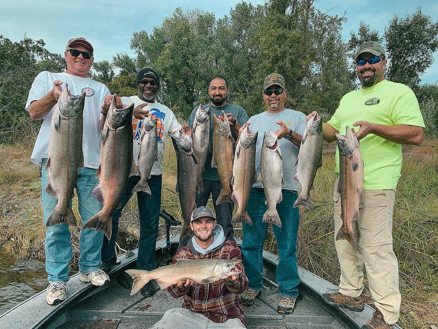 Group of six men holding large fish on a boat in a natural outdoor setting with trees in the background, some men are smiling and wearing casual outdoor clothing.
