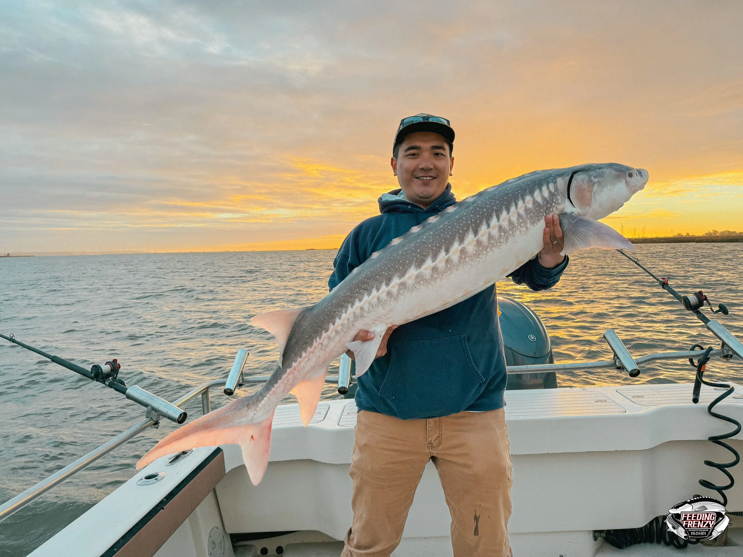 A man holding a large fish on a boat during sunset, with fishing rods in the background.