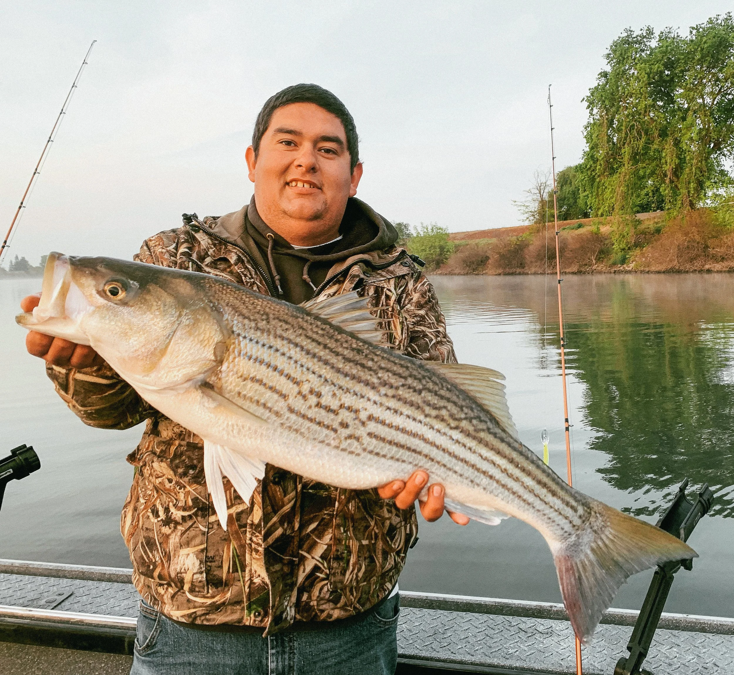 Man holding a large striped bass fish on a boat with a fishing rod and water in the background.