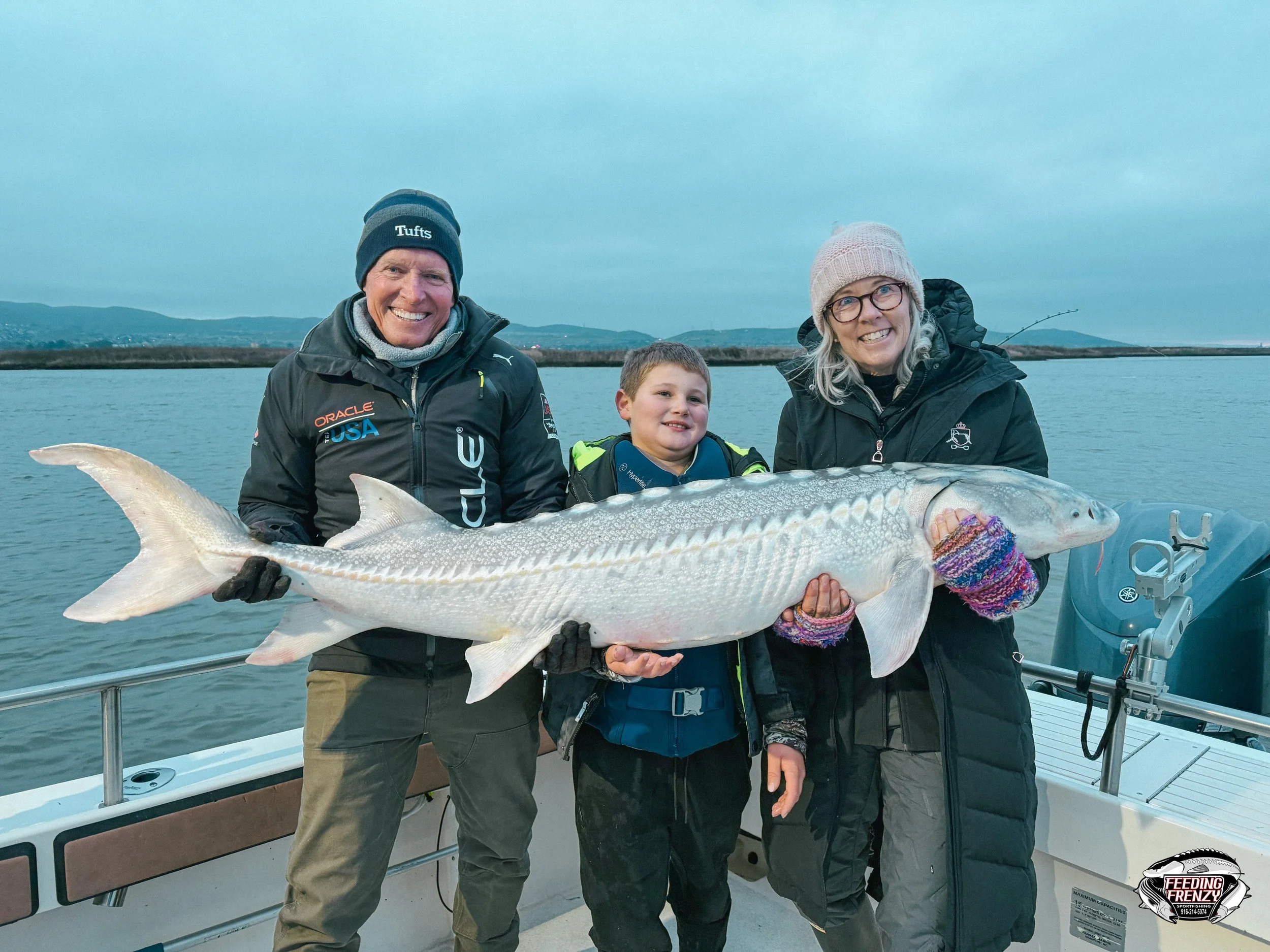 A family on a boat holding a large fish, with water and a distant shoreline in the background. The family includes an older man, a young boy, and an older woman, all smiling.