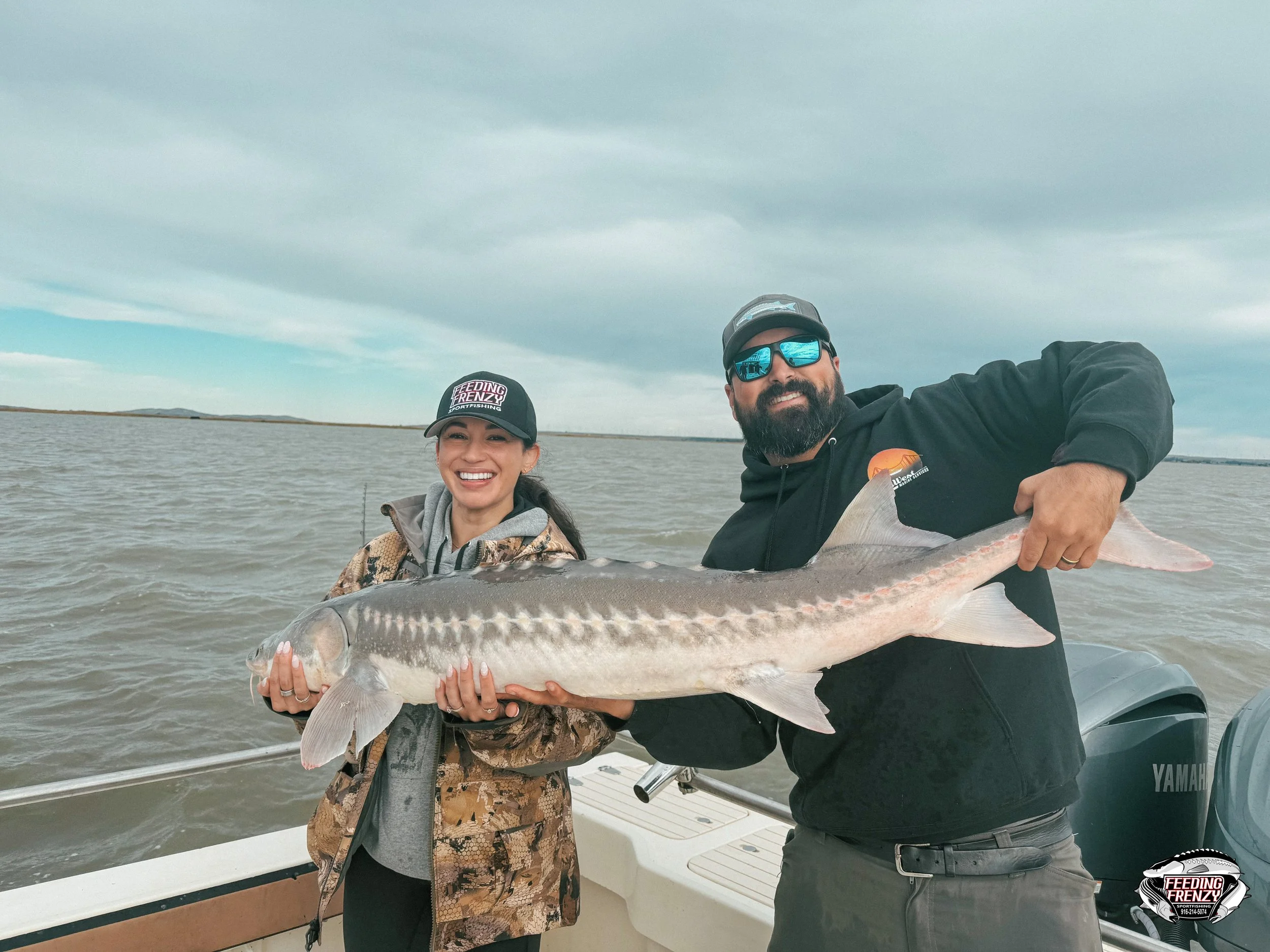 A smiling woman and man on a boat holding a large fish they caught, with water and cloudy sky in the background.