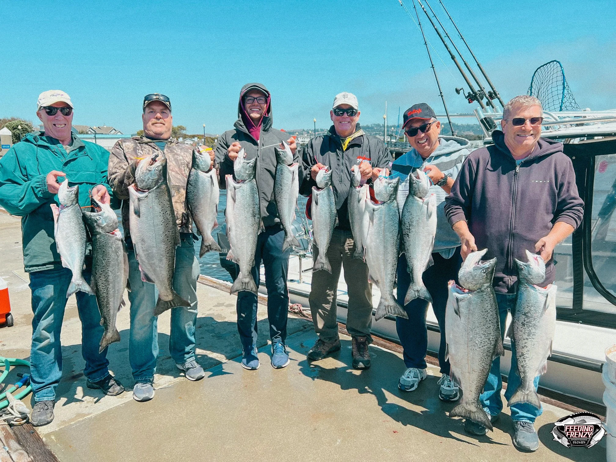 A group of seven men standing on a dock holding large fish they caught, with a boat and fishing rods in the background.