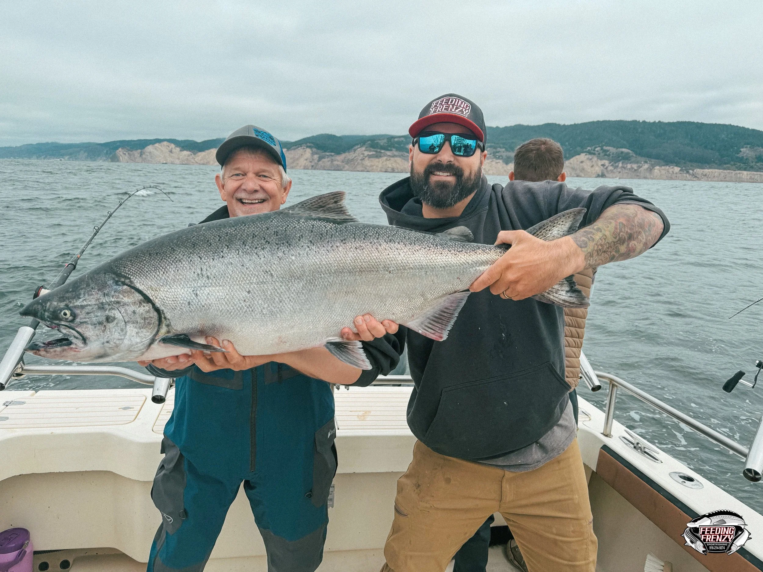 Two men on a boat holding a large fish, smiling, with a lake and distant hills in the background.