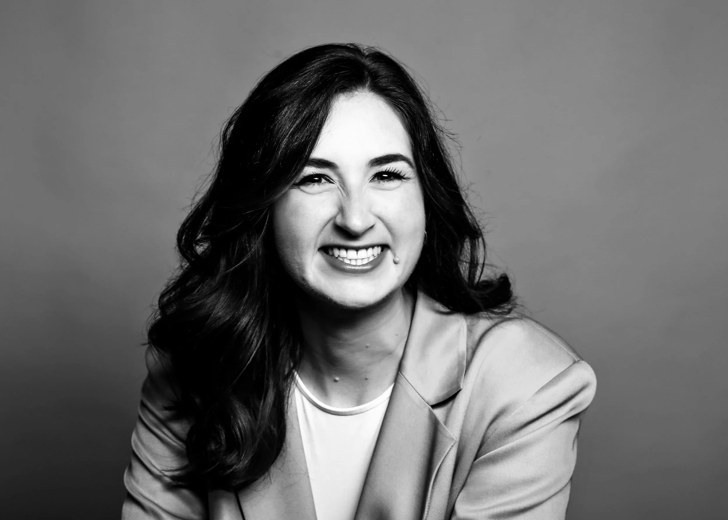 Black and white portrait of a woman with long wavy hair, smiling with teeth showing, wearing a blazer over a light top, against a plain background.