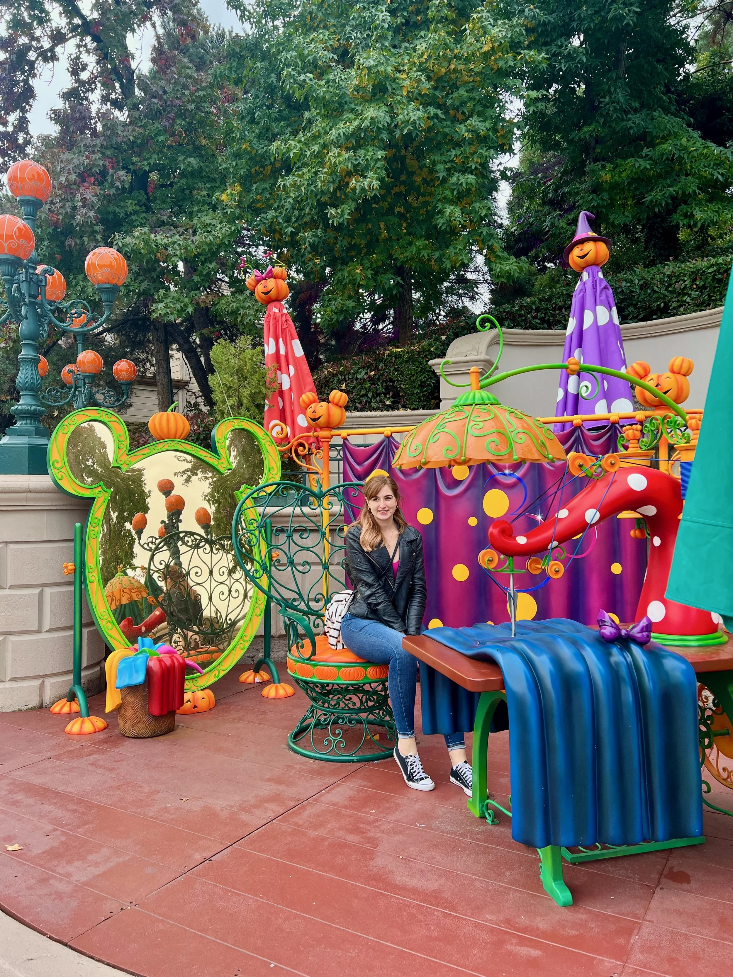 A woman sitting on a decorative wrought iron chair in front of a colorful Halloween-themed display with pumpkins, umbrellas, and a mirror with a green Mickey Mouse shaped frame.
