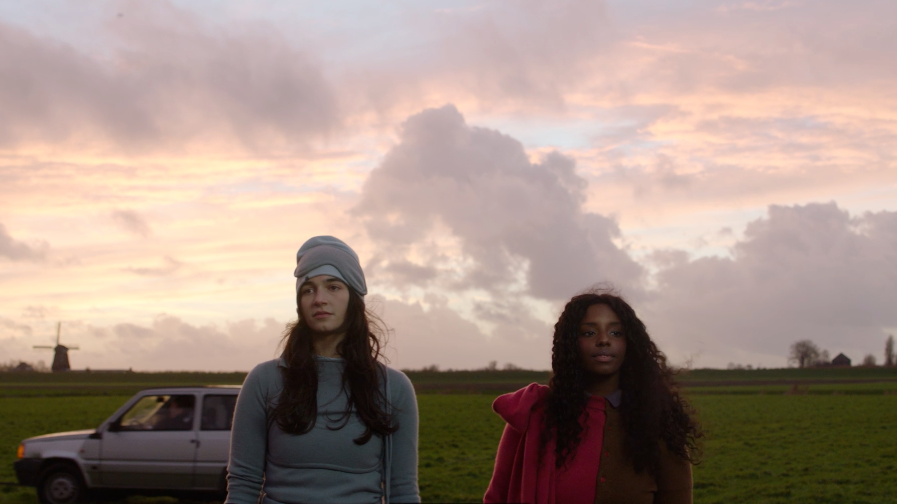 Two young women standing outdoors in a rural landscape during sunset, with a windmill and a car in the background.
