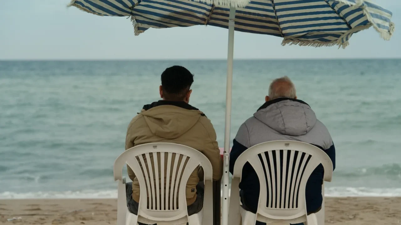 Two men sit on beach chairs under an umbrella, facing the ocean.