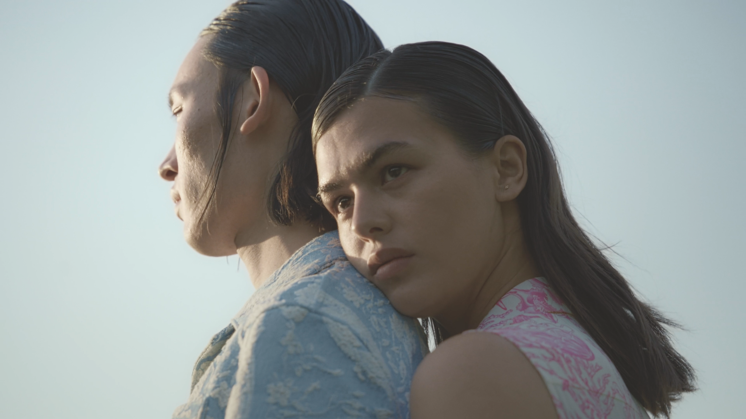 Two women with wet hair are hugging each other, one resting her head on the other's shoulder. Both appear to be outdoors with a clear sky in the background.