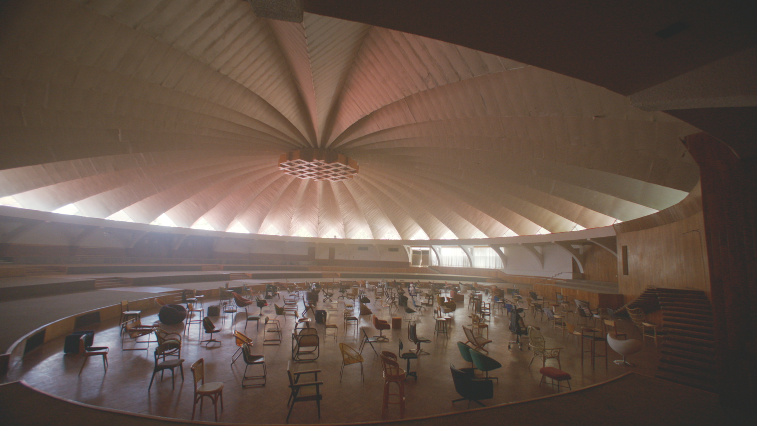 Empty theater or auditorium with numerous chairs arranged on a curved stage, under a large, dome-shaped, wooden ceiling with a radial pattern.