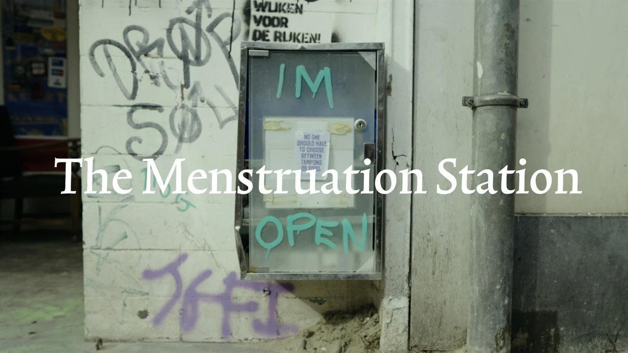 A small vending machine affixed to a graffiti-covered wall, with a sign reading 'IM OPEN' in turquoise letters. The side of the machine has a notice about tampon choosing options. The scene appears to be in an urban, possibly underground or basement setting.