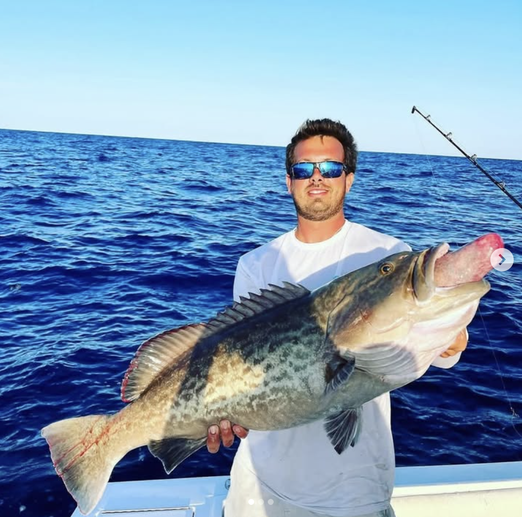 A man wearing sunglasses and a white shirt holding a large fish on a boat in the ocean.
