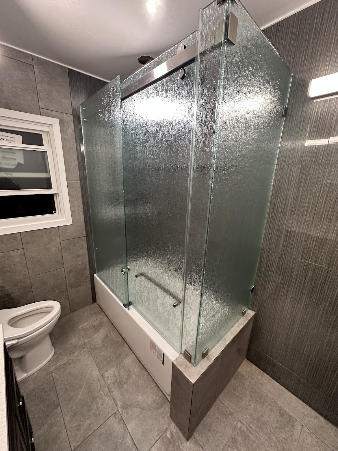 Bathroom with a glass shower enclosure, a toilet, gray tile walls, and a small window.