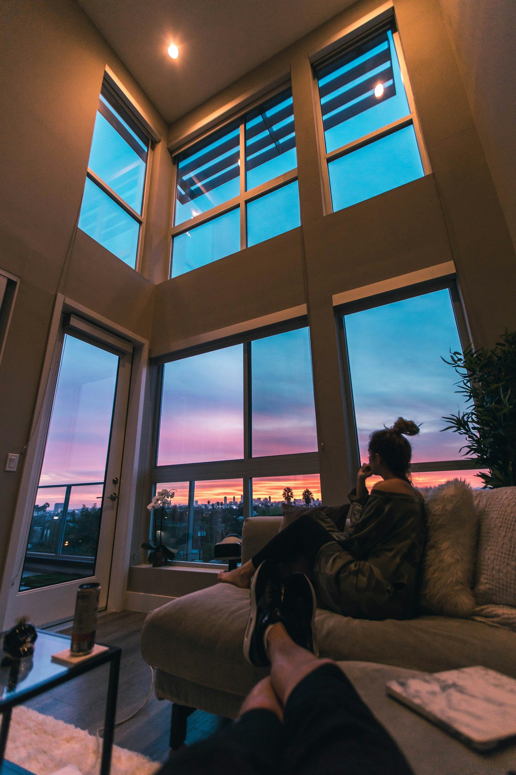 Interior of a modern living room with large floor-to-ceiling windows showing a colorful sunset sky, a woman sitting on a beige sofa looking outside, and a person reclining with legs crossed in the foreground.