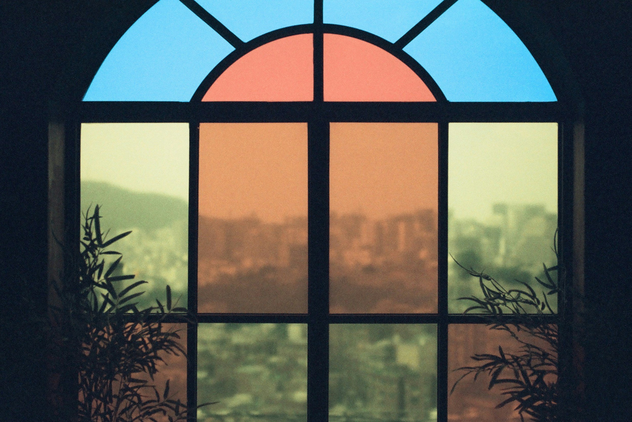 Colorful stained glass window with blue, orange, and green sections, showing a cityscape and mountains outside.