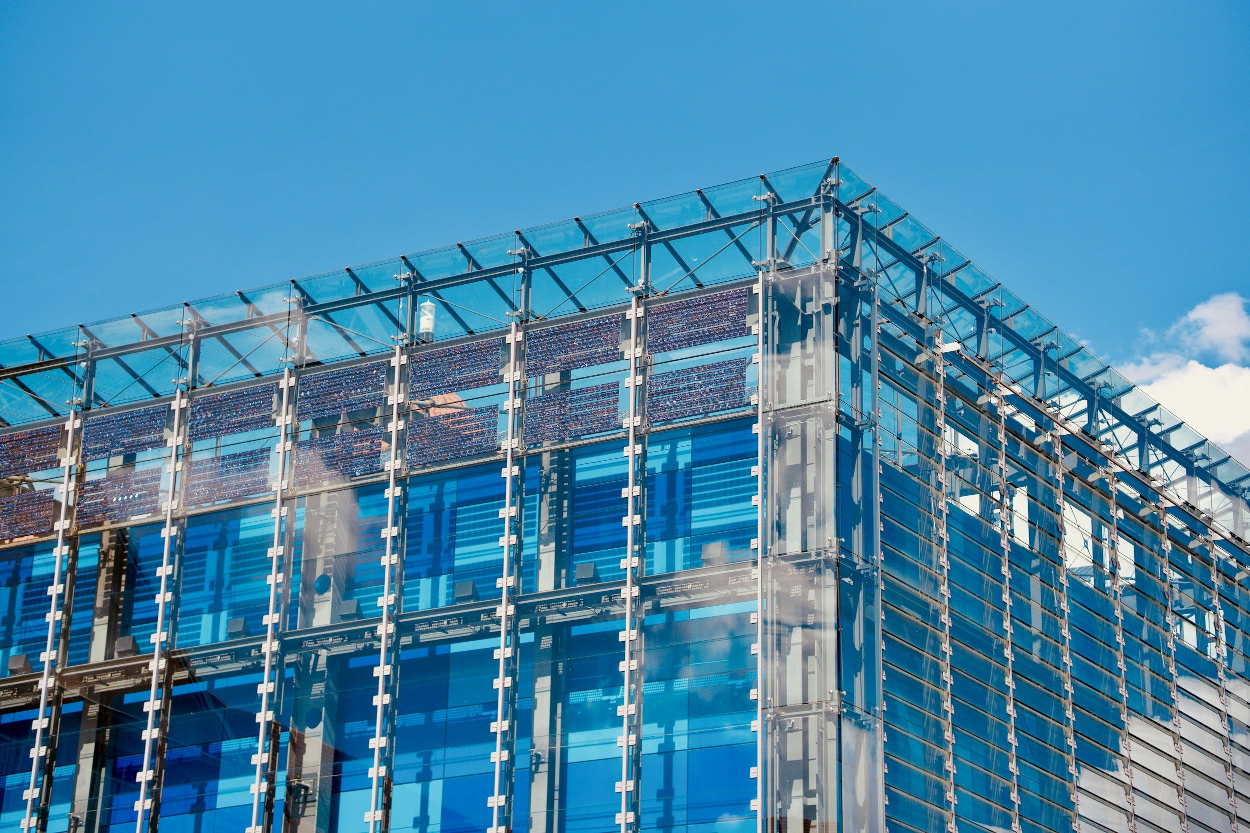 Close-up of a modern glass building with reflective blue surfaces and metal framework against a blue sky with few clouds.