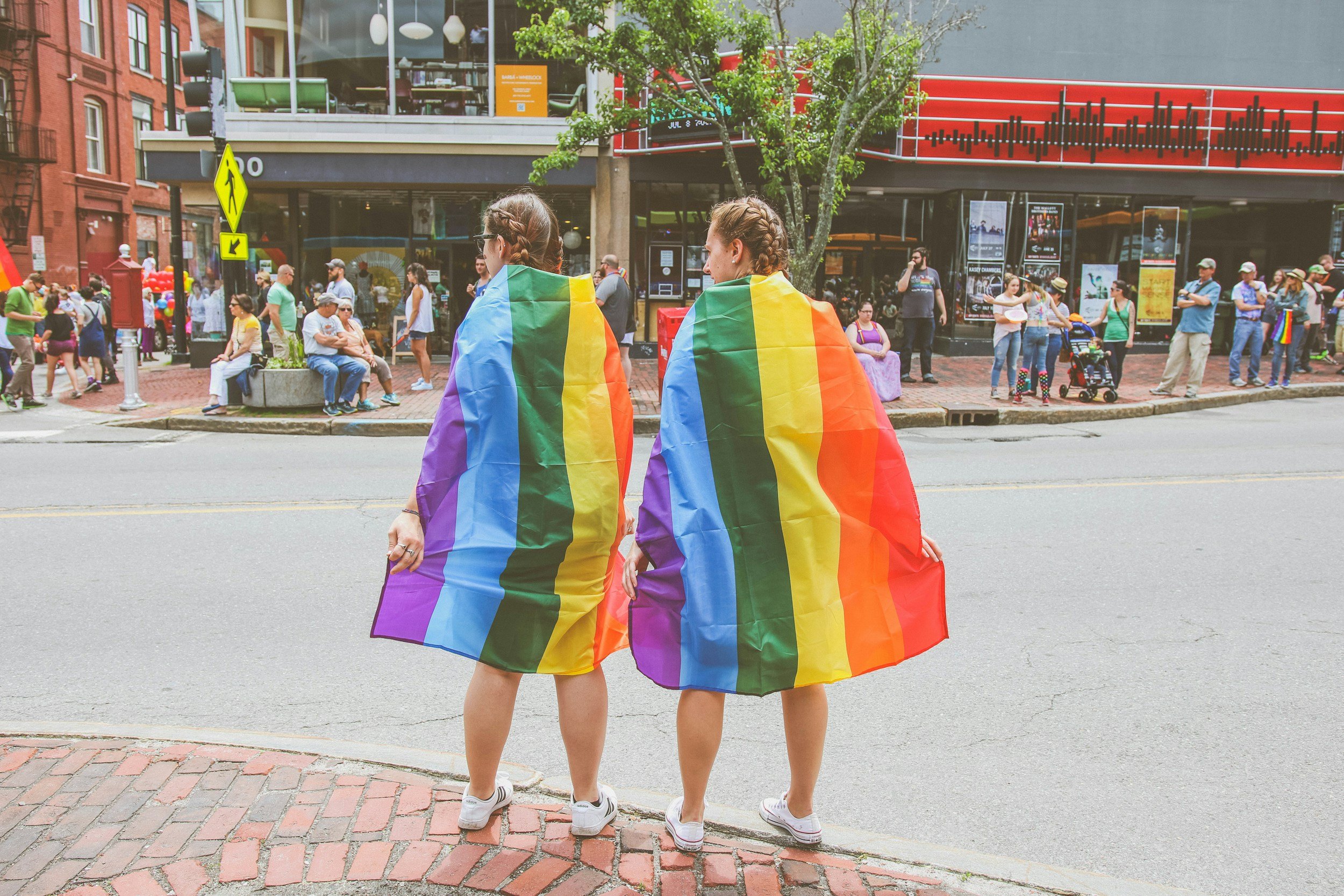 Two people on the sidewalk with rainbow flags worn as capes