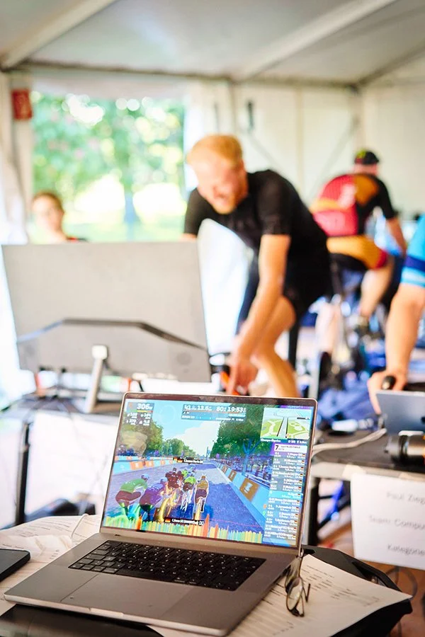 Laptop zeigt ein virtuelles Fahrradrennen, im Hintergrund Menschen im Zelt bei einer Radveranstaltung.