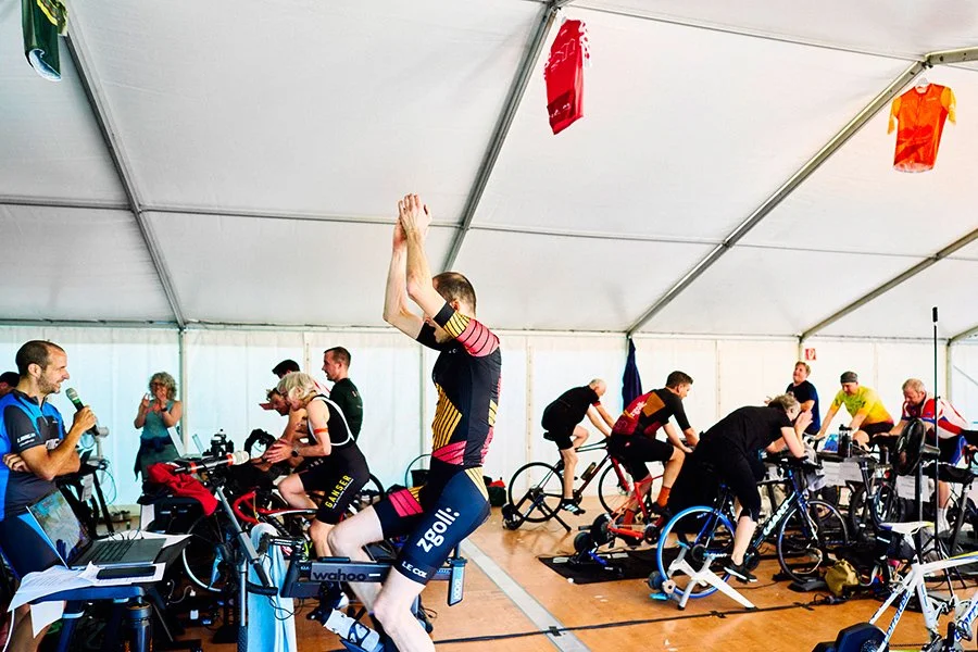 Menschen trainieren indoor auf Spinner- und Rennrädern, einige sitzen, andere stehen oder dehnen sich. Es sind Sportler, die für Radrennen oder -training vorbereitet werden.