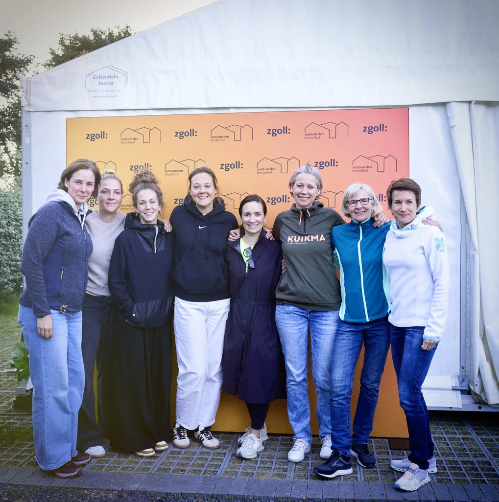 Sieben Frauen stehen nebeneinander vor einem orangefarbenen Hintergrund mit Text und einem Logo, sie sind lächelnd und haben die Arme umeinander gelegt.