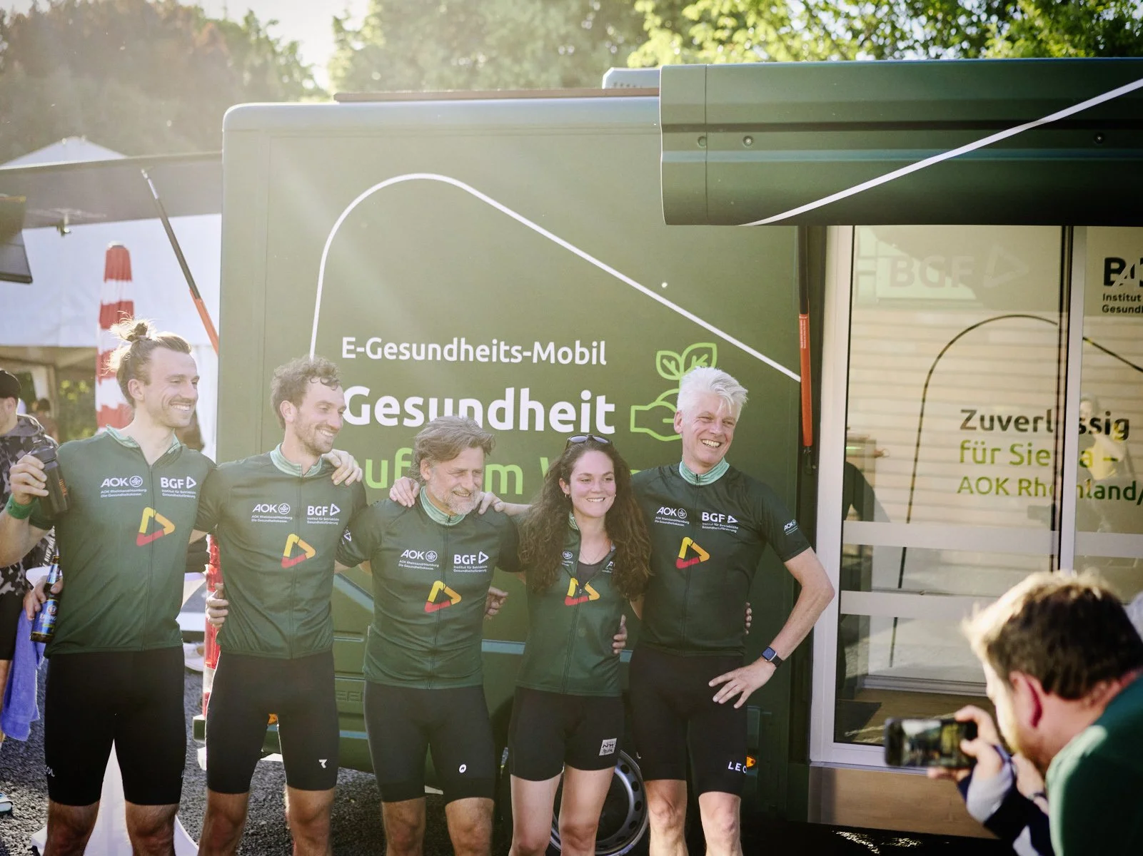 Fünf Menschen in Sportkleidung posieren lachend vor einem grünen E-Gesundheits-Mobil, während im Vordergrund eine Person ein Foto macht.