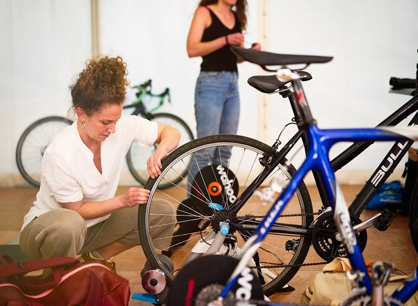 Zwei Frauen bereiten Fahrräder für eine Fahrt vor, eine repariert ein Fahrrad im Vordergrund, während die andere im Hintergrund ein weiteres Fahrrad hält.