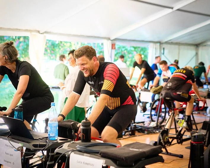 Menschen beim Indoor-Spinning-Workout in einer Zeltanlage, lachende Teilnehmer beim Training, verschiedene Fahrräder, Trainerin, Wasserflaschen, Trainingsgeräte.