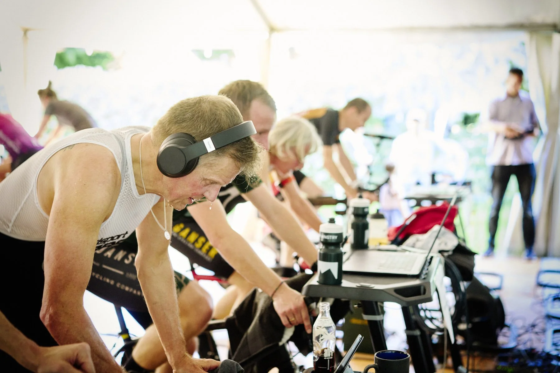 Leute beim Indoor-Cycling-Training, einige tragen Kopfhörer und trinken Wasser, während sie auf Fahrrädern trainieren.