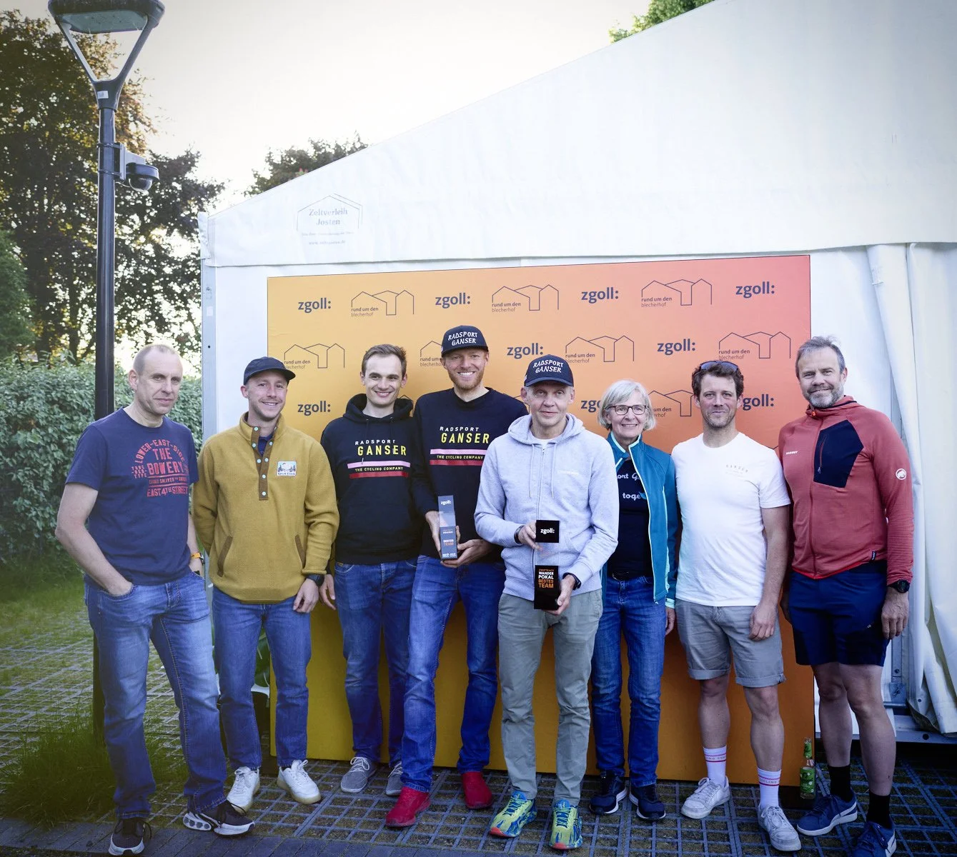 Eine Gruppe von acht Menschen steht vor einem orangefarbenen Hintergrund mit Logos und Text, hält Trophäen und feiert einen Erfolg bei einem Radsport-Event.