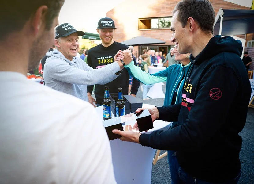 Männer bei einer Feier, einer reicht dem anderen ein Geschenk, im Hintergrund Getränke und Menschen bei einer Outdoor-Veranstaltung.