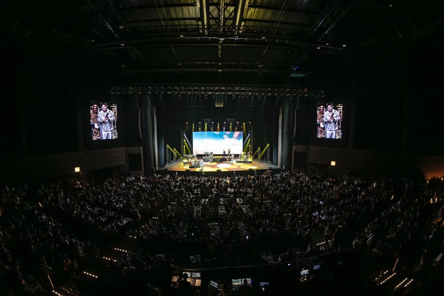 Show de música em um palco grande com iluminação colorida, público assistindo sentado em poltronas escurecidas, duas telas de vídeo laterais e uma tela grande ao fundo.