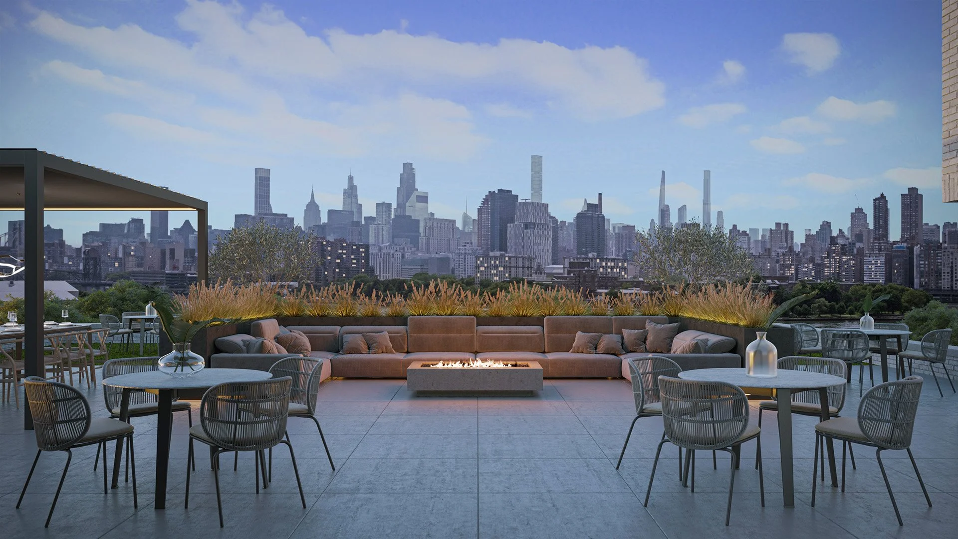 Rooftop outdoor lounge area with a view of a city skyline, featuring a sofa with pillows, tables with vases, and a fire pit in the center.