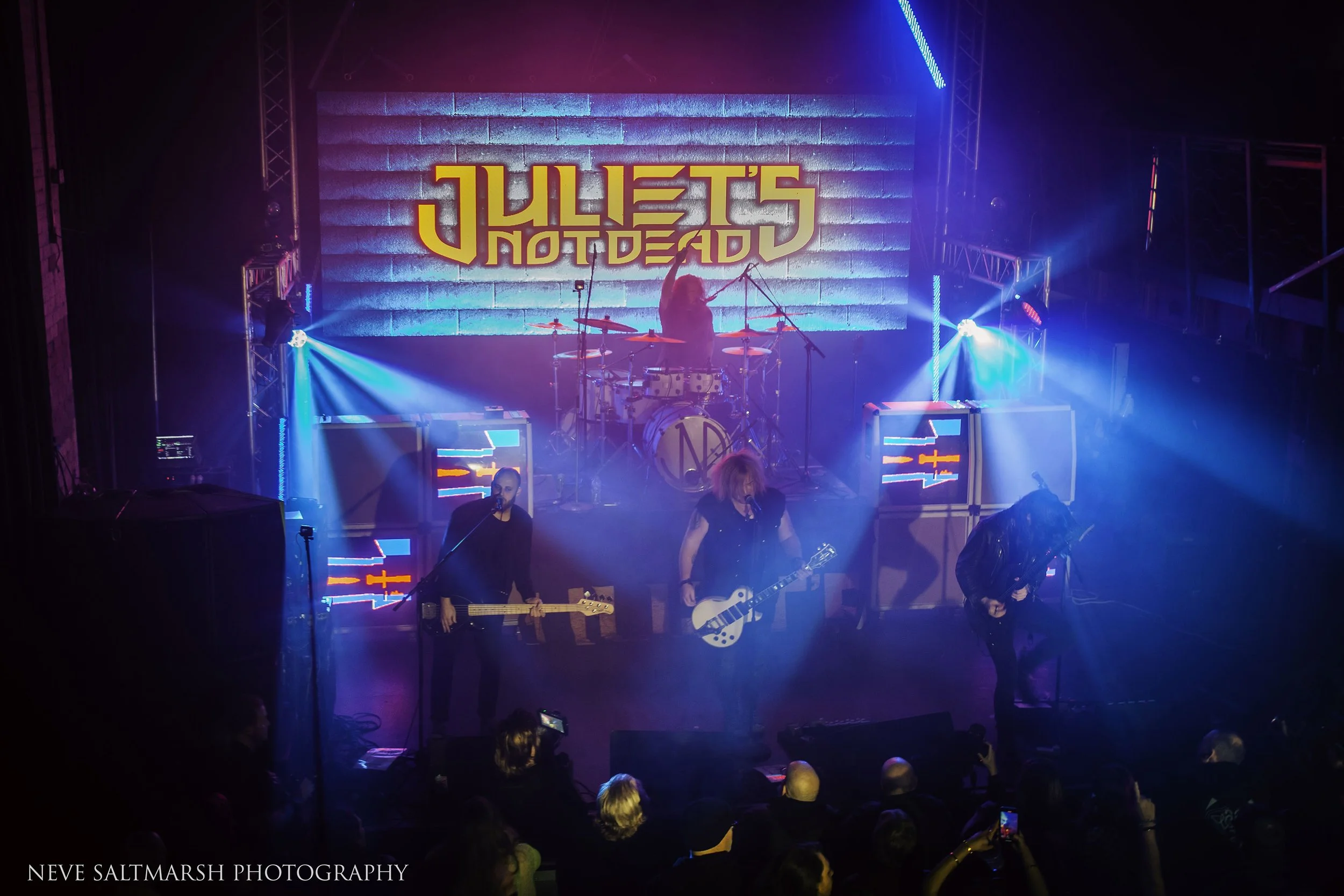 A band performing on stage with a neon sign that reads 'JULIET'S NOT DEAD' in the background, colored stage lights, and an audience watching.