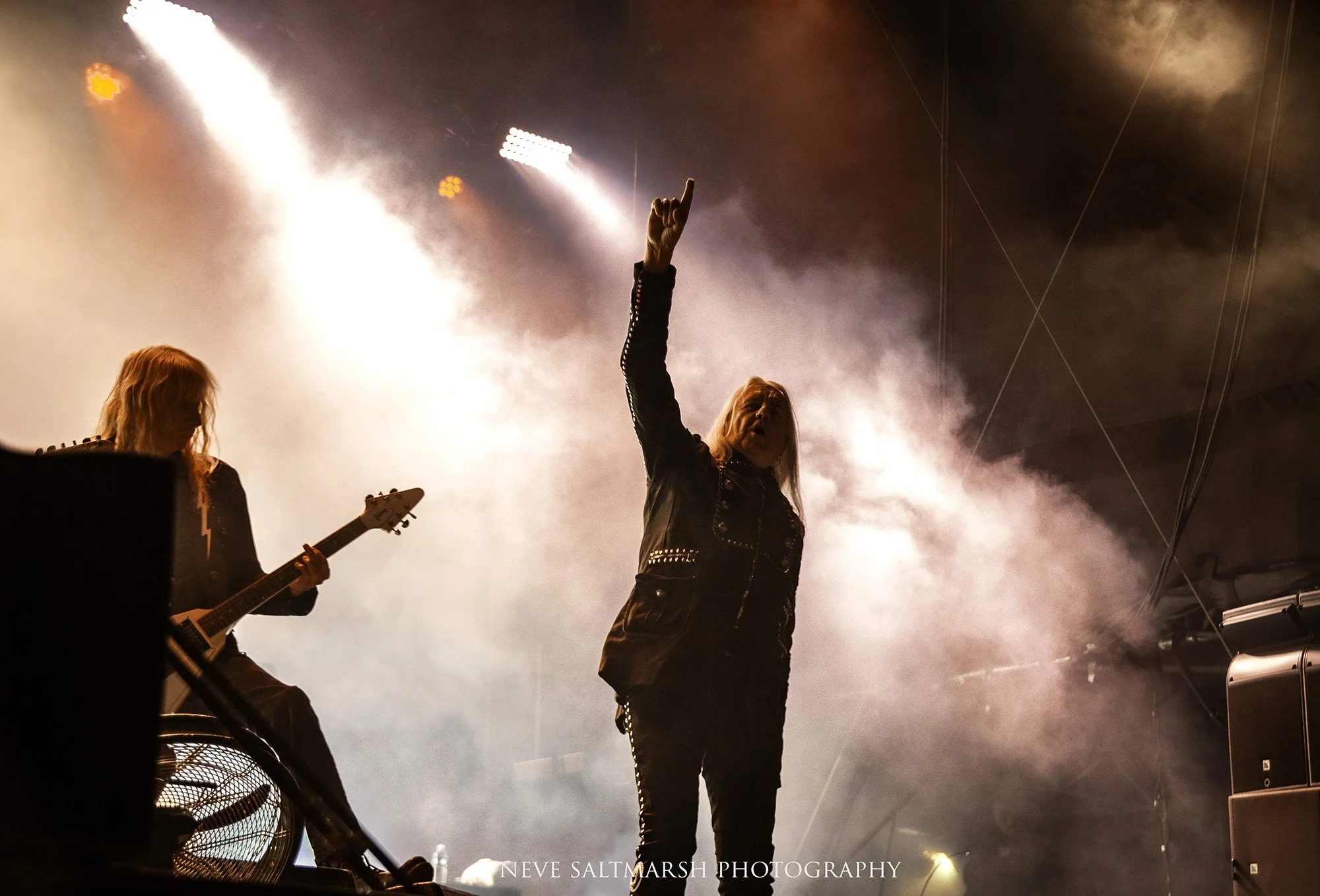 A male musician on stage raising his right hand with index finger pointing upward, surrounded by smoke and bright stage lights, with a guitarist on his left playing an electric guitar.