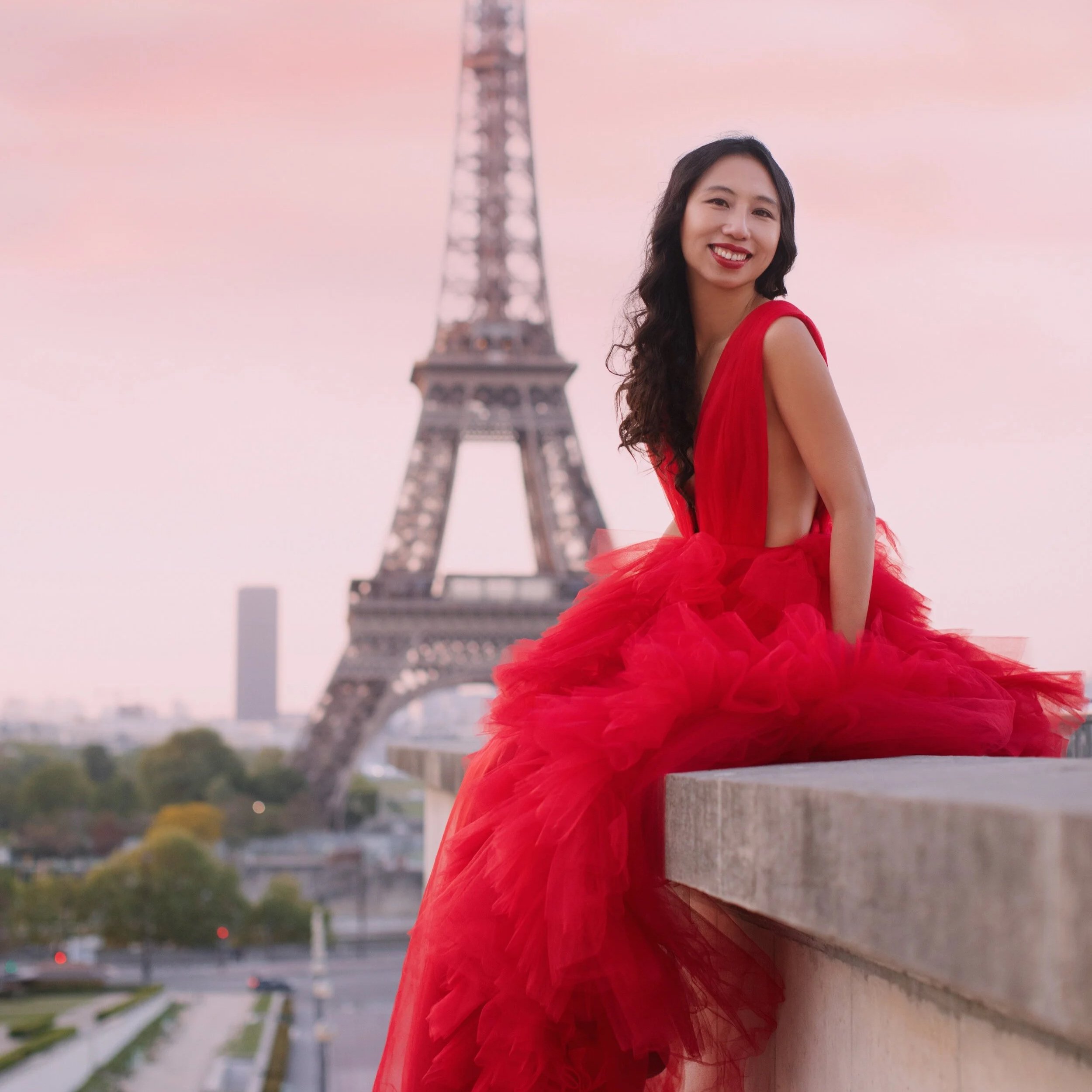 A woman in a red gown sitting on a ledge with the Eiffel Tower in the background during sunset in Paris.
