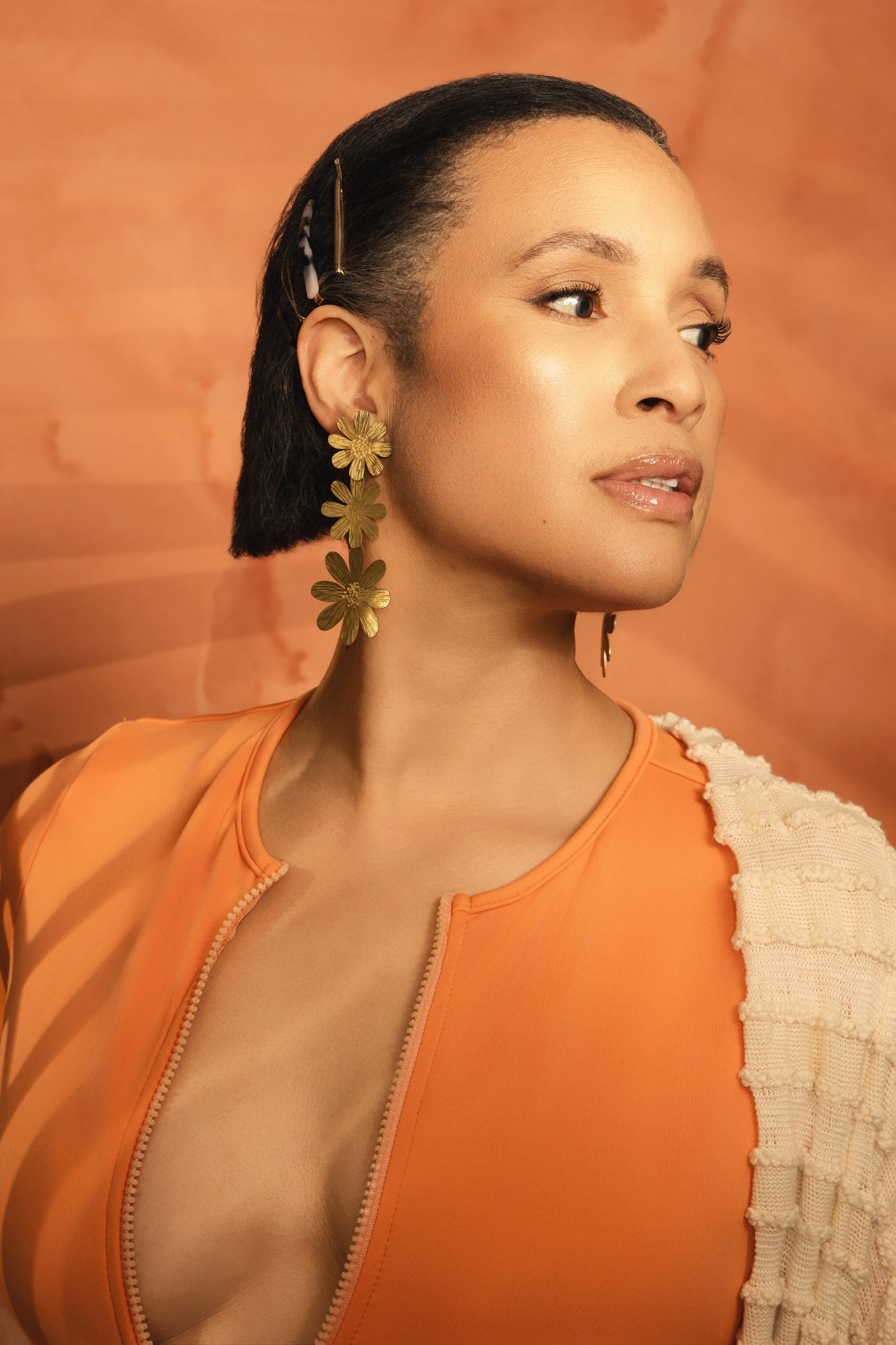 A woman with dark, straight, short hair wearing large gold flower earrings and orange clothing with textured cream fabric on the right shoulder, posed against a warm gradient background. Swimsuit fashion photographer