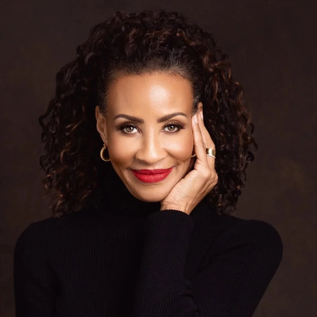 Black and white photo of a woman with dark hair, wearing lace lingerie and a layered necklace, lying on a bed.