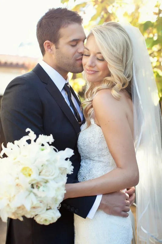 A newlywed couple, a man and a woman, sharing an intimate moment outdoors. The man is in a black suit with a white shirt and striped tie, gently holding the bride from behind. The bride is in a white lace wedding dress with a veil, smiling with her e