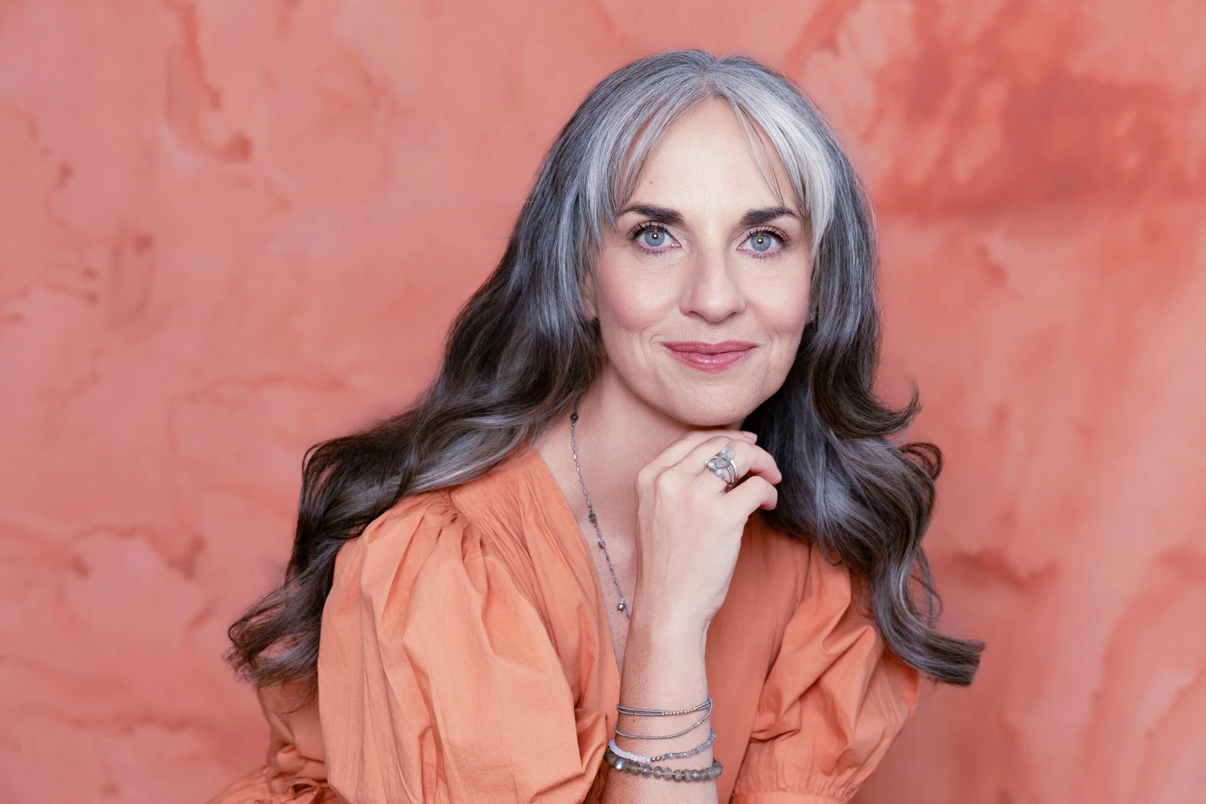 A woman with gray and dark brown hair, blue eyes, wearing an orange top, smiling softly, standing in front of a peach-colored textured background.