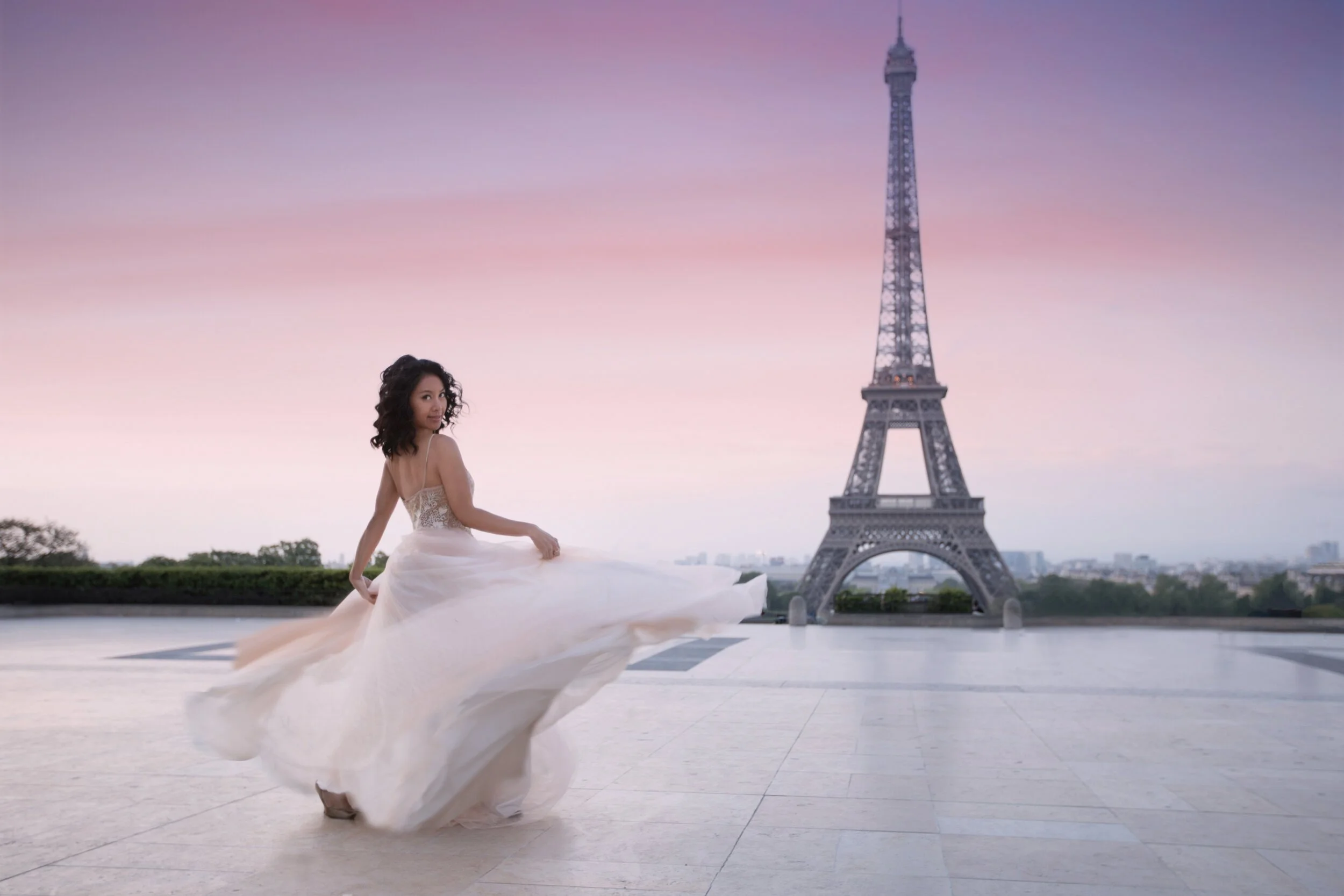 A woman in a white wedding dress poses in front of the Eiffel Tower during sunset in Paris. Destination Paris portrait photographer