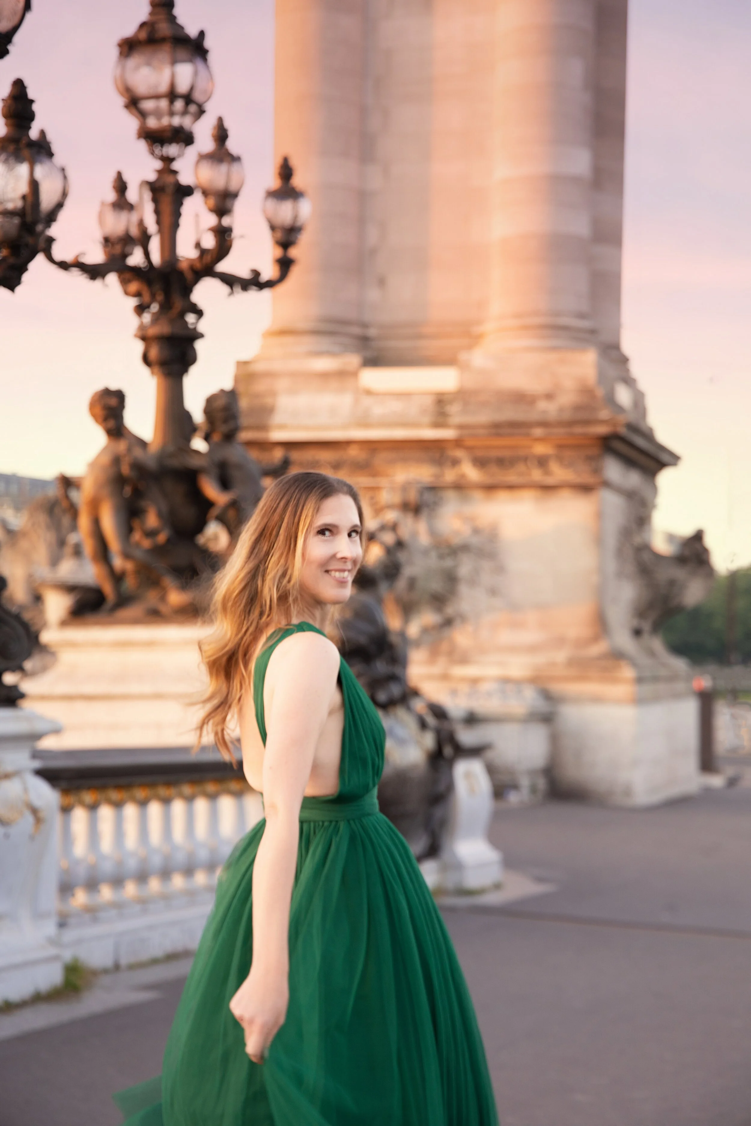 A woman in a flowing green dress smiling and looking at the camera, standing outdoors near a historic monument with sculptures and ornate street lamps, during sunset. Destination Paris portrait photographer