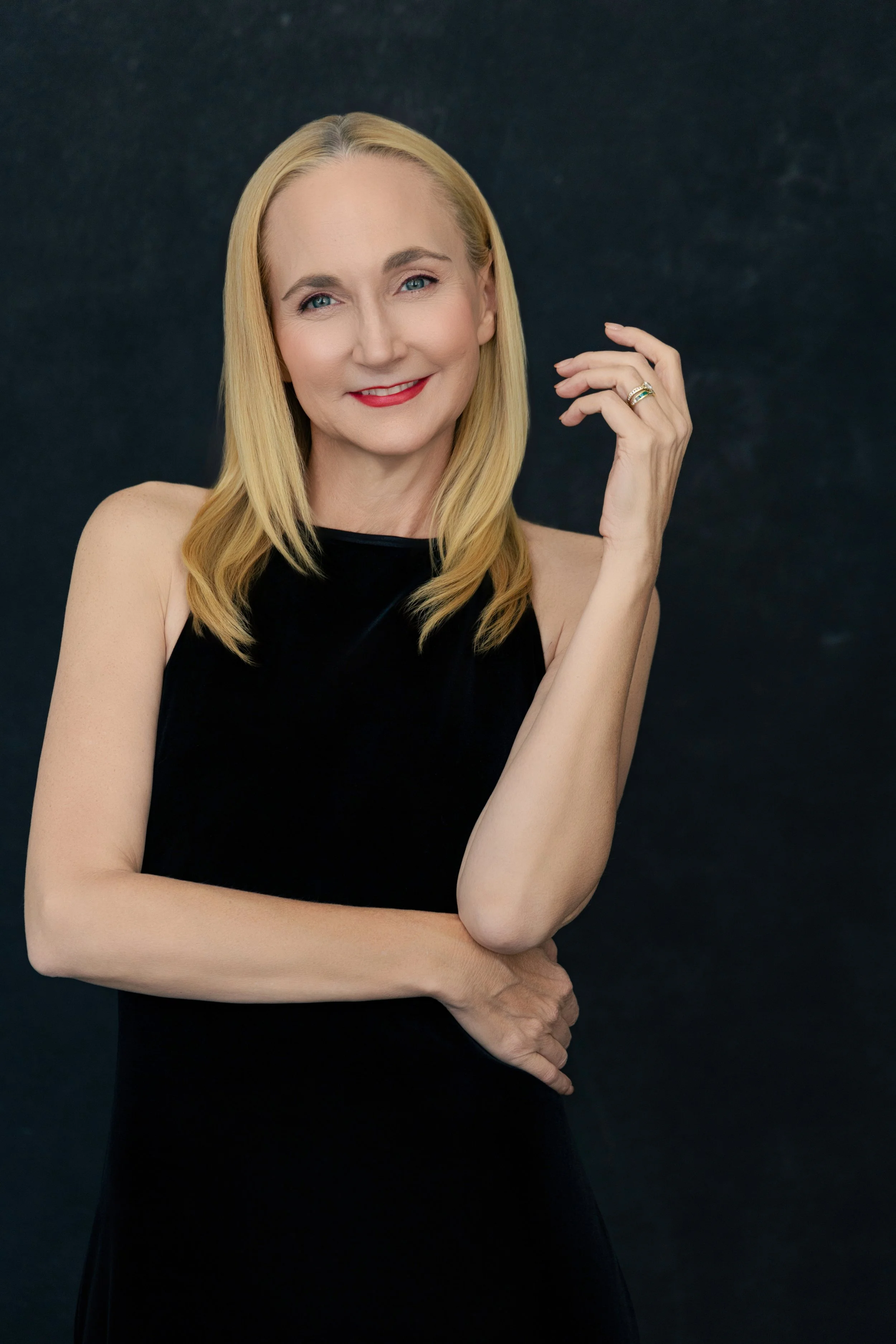 A woman with blonde hair wearing a sleeveless black dress, smiling, with one hand touching her face and the other arm crossed, against a dark background.