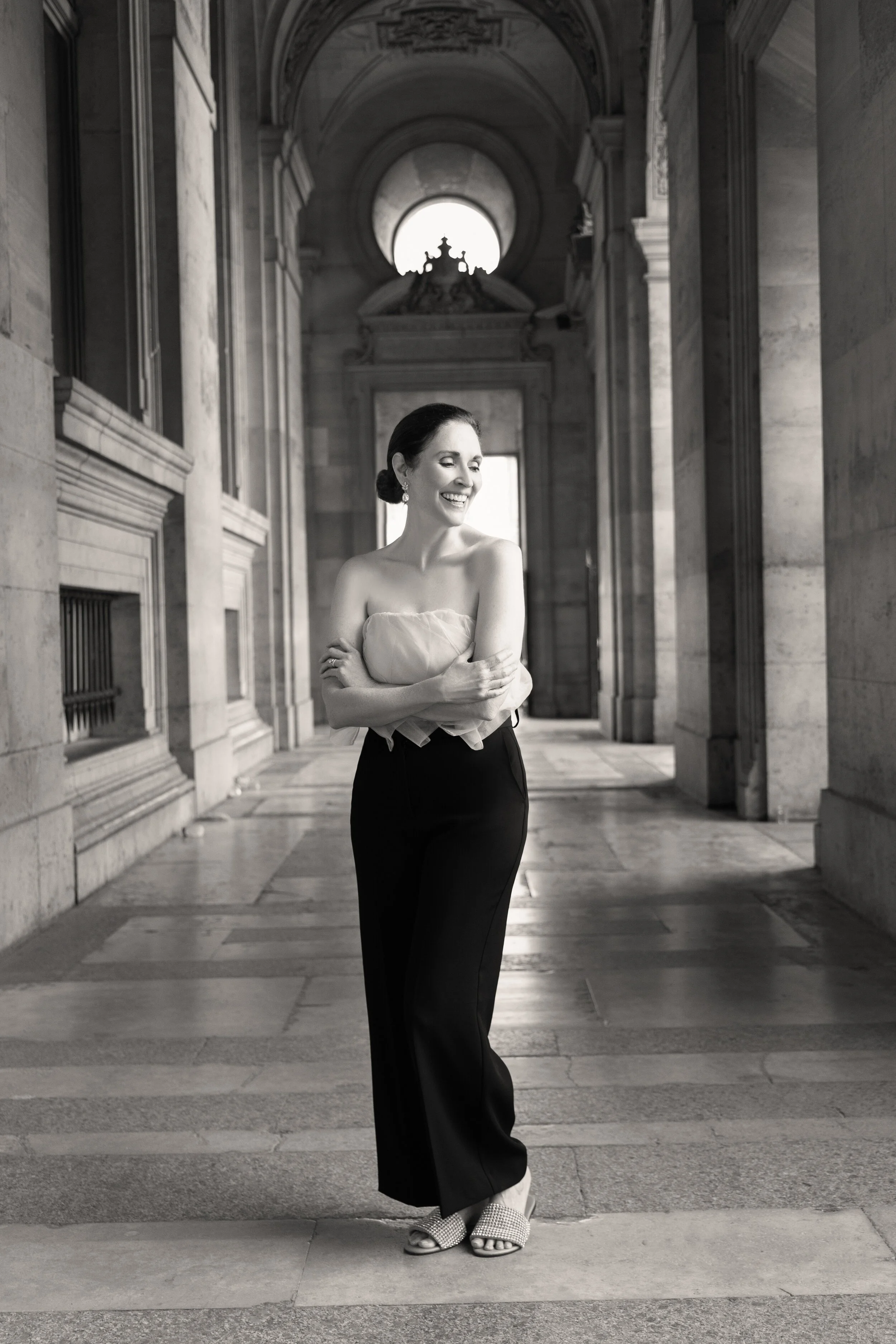 A woman with dark hair tied back, smiling, standing in an elegant, classical building with arches and high ceilings, dressed in a strapless top, black pants, and open-toed heels.