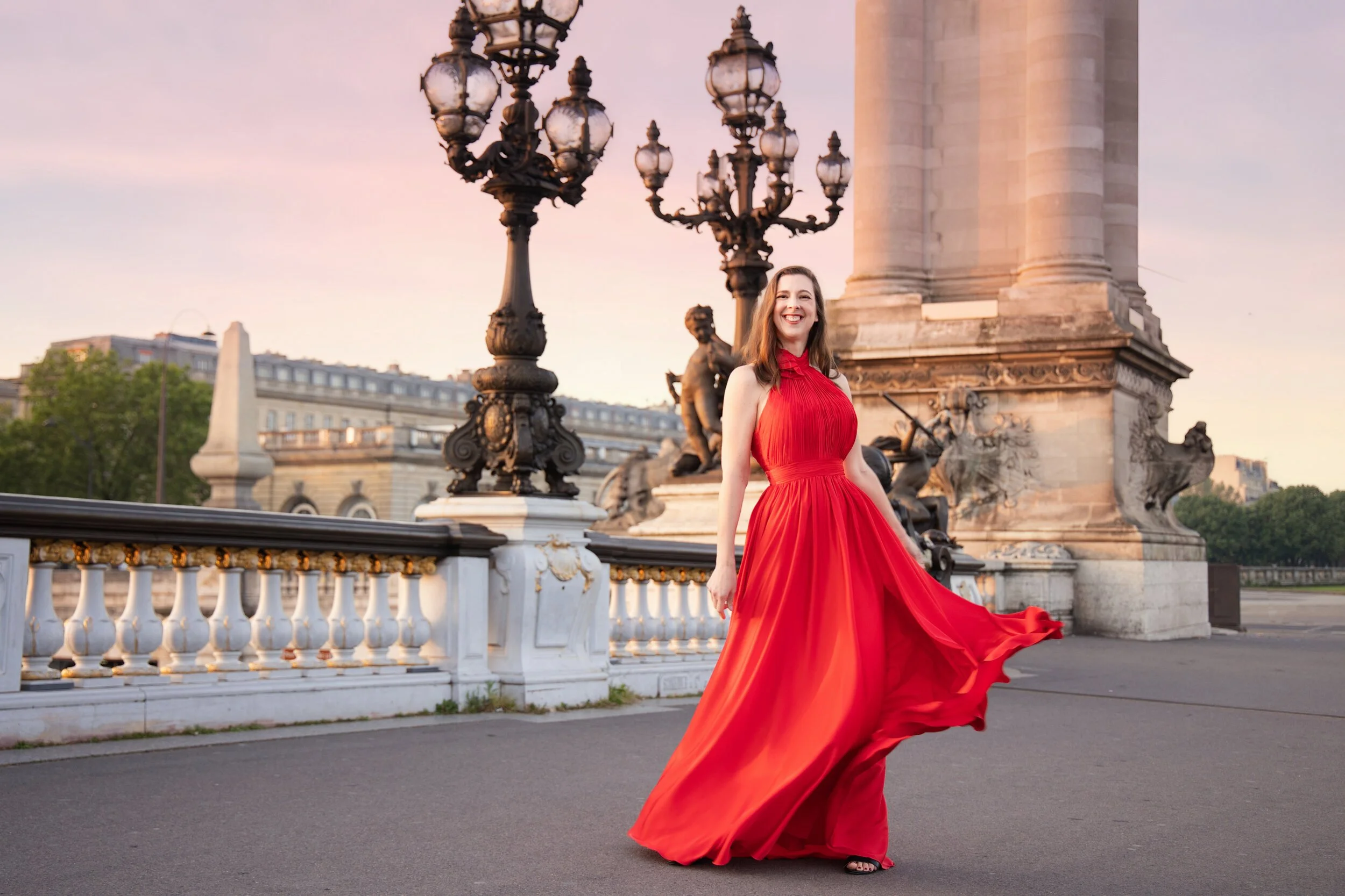 A woman in a flowing red dress standing on a bridge at sunset, smiling, with ornate street lamps and a monument in the background.