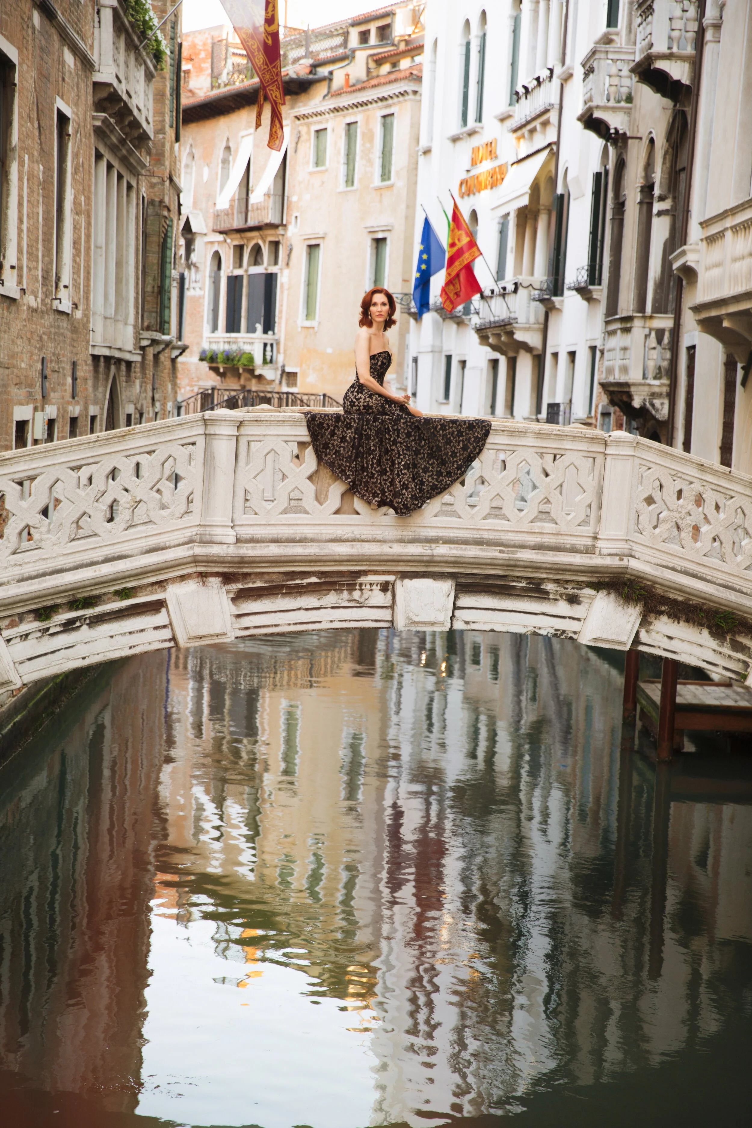 A woman in a black and gold dress sitting on a small bridge over a canal in Venice, Italy, with European and Venetian flags in the background. Destination Paris portrait photographer