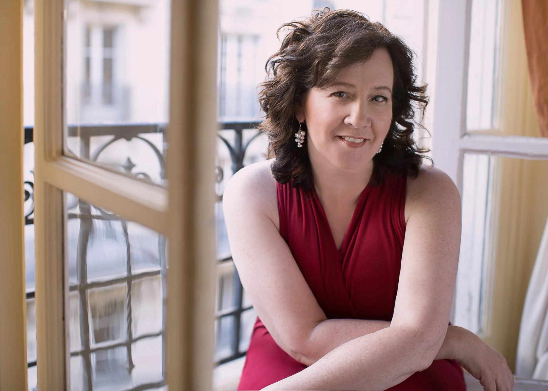 A woman with dark curly hair, wearing a red sleeveless dress and pearl earrings, sitting near a window with decorative wrought iron balcony, smiling at the camera.